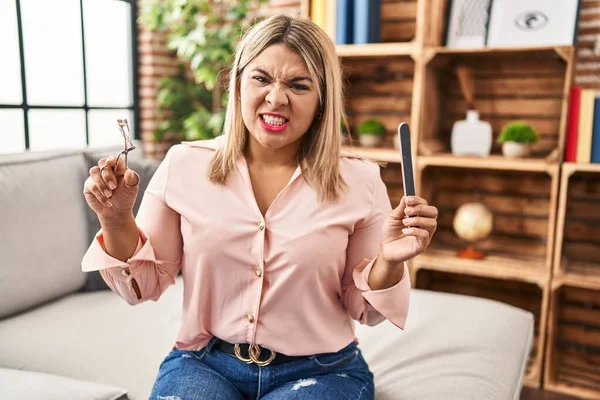 Young hispanic woman holding eye lashes curler and nail file angry and mad screaming frustrated and furious, shouting with anger. rage and aggressive concept.