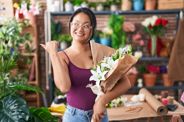Asian young woman at florist shop holding bouquet of flowers pointing thumb up to the side smiling happy with open mouth