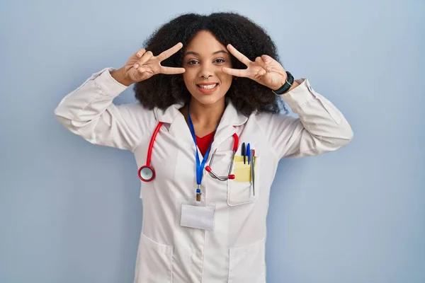 顔の上に指で平和のシンボルを行う医師の制服と聴診器を身に着けている若いアフリカ系アメリカ人女性 勝利を示す明るい笑顔 — ストック写真