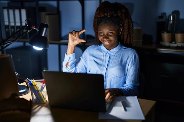African woman working at the office at night smiling and confident gesturing with hand doing small size sign with fingers looking and the camera. measure concept.