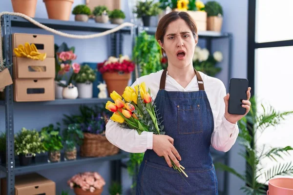 Brunette woman working at florist shop holding smartphone in shock face, looking skeptical and sarcastic, surprised with open mouth