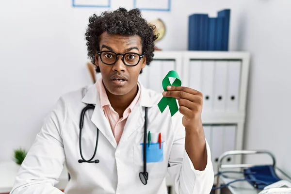 African doctor man holding support green ribbon scared and amazed with open mouth for surprise, disbelief face