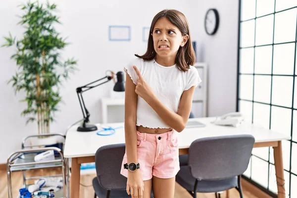Young hispanic girl standing at pediatrician clinic pointing aside worried and nervous with forefinger, concerned and surprised expression 