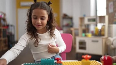 Adorable hispanic girl playing with construction blocks sitting on table at classroom