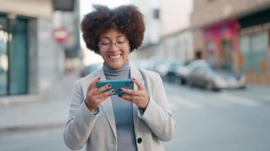 Afro-Amerikalı kadın gülümsüyor. Sokakta akıllı telefondan videolar izliyor.