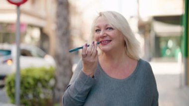 Orta yaşlı, sarışın, gülümseyen, kendine güvenen bir kadın caddede akıllı telefondan konuşuyor.