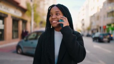 Genç Afrikalı Amerikalı kadın gülümsüyor. Kendine güveni tam. Sokakta akıllı telefondan konuşuyor.