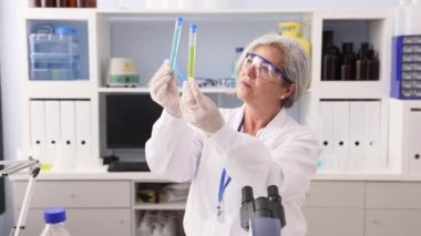 Orta yaşlı, gri saçlı, bilim adamı üniforması giyen bir kadın laboratuarda test tüplerini karşılaştırıyor.