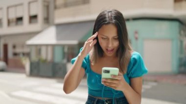 Young hispanic woman using smartphone with surprise expression at street