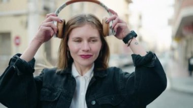Genç beyaz kadın, kendine güvenen bir şekilde gülümsüyor müzik dinliyor ve sokakta dans ediyor.