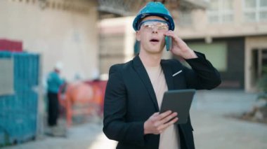 Young hispanic man architect talking on the smartphone using touchpad at street