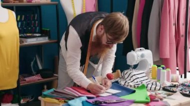Young redhead man tailor measuring t shirt writing on notebook at clothing factory