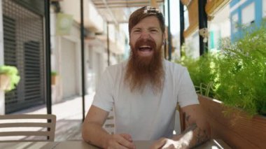 Young redhead man smiling confident sitting on table at coffee shop terrace