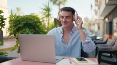 Young hispanic man listening to music sitting on table at coffee shop terrace