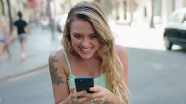Young blonde woman using smartphone with cheerful expression at street