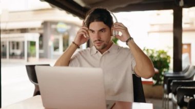 Young hispanic man using laptop and headphones at coffee shop terrace
