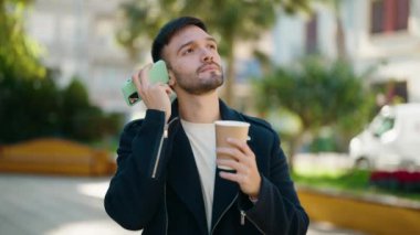 Young hispanic man drinking coffee listening audio message by the smartphone at park