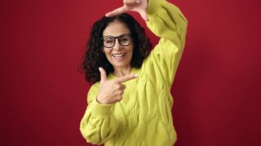 Middle age hispanic woman smiling confident doing photo gesture with hands over isolated red background