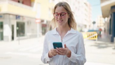 Young blonde woman smiling and using smartphone at street