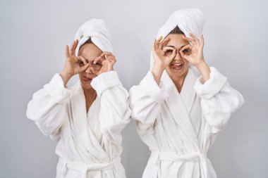 Middle age woman and daughter wearing white bathrobe and towel doing ok gesture like binoculars sticking tongue out, eyes looking through fingers. crazy expression. 