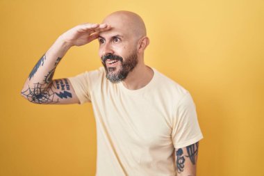 Hispanic man with tattoos standing over yellow background very happy and smiling looking far away with hand over head. searching concept. 