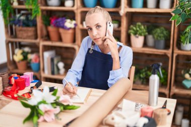 Çiçekçide, çiçekçi, çiçekçi defterine akıllı telefon yazısıyla konuşan genç sarışın kadın.
