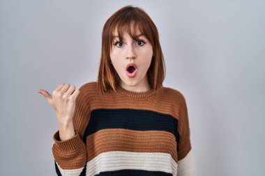 Young beautiful woman wearing striped sweater over isolated background surprised pointing with hand finger to the side, open mouth amazed expression. 