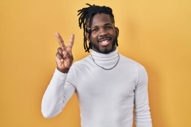 Sarı arka plan üzerine balıkçı yaka kazak giyen Afrikalı bir adam iki numaralı parmaklarını gösterirken kendinden emin ve mutlu bir şekilde gülümsüyor.. 