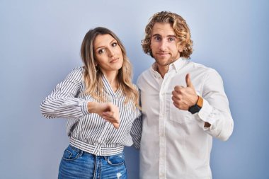 Young couple standing over blue background doing thumbs up and down, disagreement and agreement expression. crazy conflict 