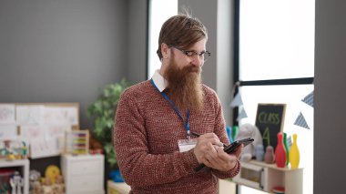 Young redhead man preschool teacher writing on touchpad standing at kindergarten