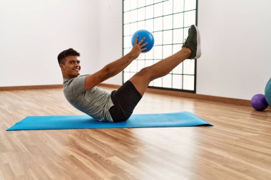 Genç İspanyol adam gülümsüyor. Spor merkezinde top kullanarak karın kasları çalıştırıyor.
