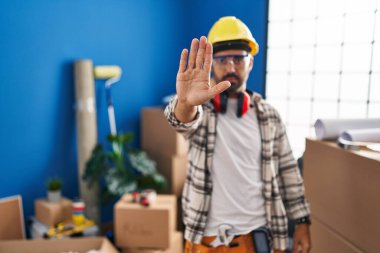 Young hispanic man with beard working at home renovation doing stop sing with palm of the hand. warning expression with negative and serious gesture on the face. 