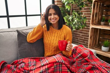 Young hispanic woman sitting on the sofa drinking a coffee at home smiling with hand over ear listening an hearing to rumor or gossip. deafness concept. 