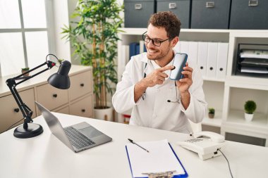 Doktor üniforması giyen genç İspanyol adam klinikte akıllı telefon olduğunu gösteren teletıp taşıyor.