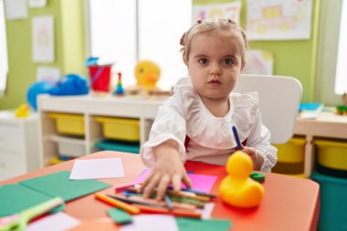 Sevimli sarışın anaokulu öğrencisi anaokulunda masaya oturmuş kağıda resim çiziyor.