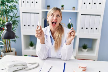 Beautiful dentist woman holding ordinary toothbrush and electric toothbrush angry and mad screaming frustrated and furious, shouting with anger looking up. 