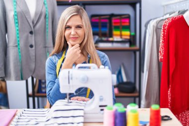 Blonde woman dressmaker designer using sew machine looking confident at the camera smiling with crossed arms and hand raised on chin. thinking positive. 