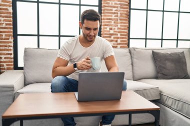 Young hispanic man using laptop and drinking coffee sitting on sofa at home