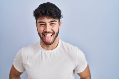 Hispanic man with beard standing over white background sticking tongue out happy with funny expression. emotion concept. 