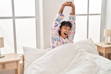 Young beautiful hispanic woman waking up stretching arms yawning at bedroom