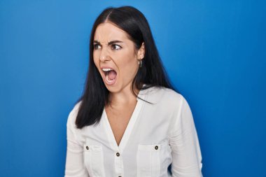 Young hispanic woman standing over blue background angry and mad screaming frustrated and furious, shouting with anger. rage and aggressive concept. 