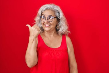 Middle age woman with grey hair standing over red background smiling with happy face looking and pointing to the side with thumb up. 
