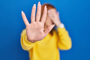 Young woman standing over blue background covering eyes with hands and doing stop gesture with sad and fear expression. embarrassed and negative concept. 