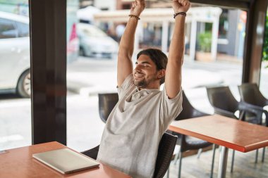 Young hispanic man stretching arms sitting on table at coffee shop terrace