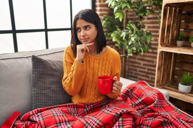 Young hispanic woman sitting on the sofa drinking a coffee at home with hand on chin thinking about question, pensive expression. smiling with thoughtful face. doubt concept. 