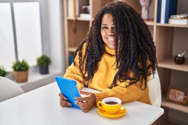 African american woman using touchpad drinking coffee sitting on table at home