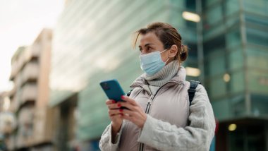 Orta yaşlı, tıbbi maske takan bir kadın sokakta akıllı telefon kullanıyor.