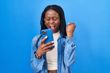 African woman with braids using smartphone typing message annoyed and frustrated shouting with anger, yelling crazy with anger and hand raised 