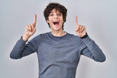Young man standing over isolated background smiling amazed and surprised and pointing up with fingers and raised arms. 