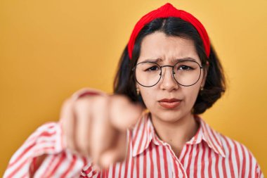 Young hispanic woman wearing glasses over yellow background pointing with finger to the camera and to you, confident gesture looking serious 